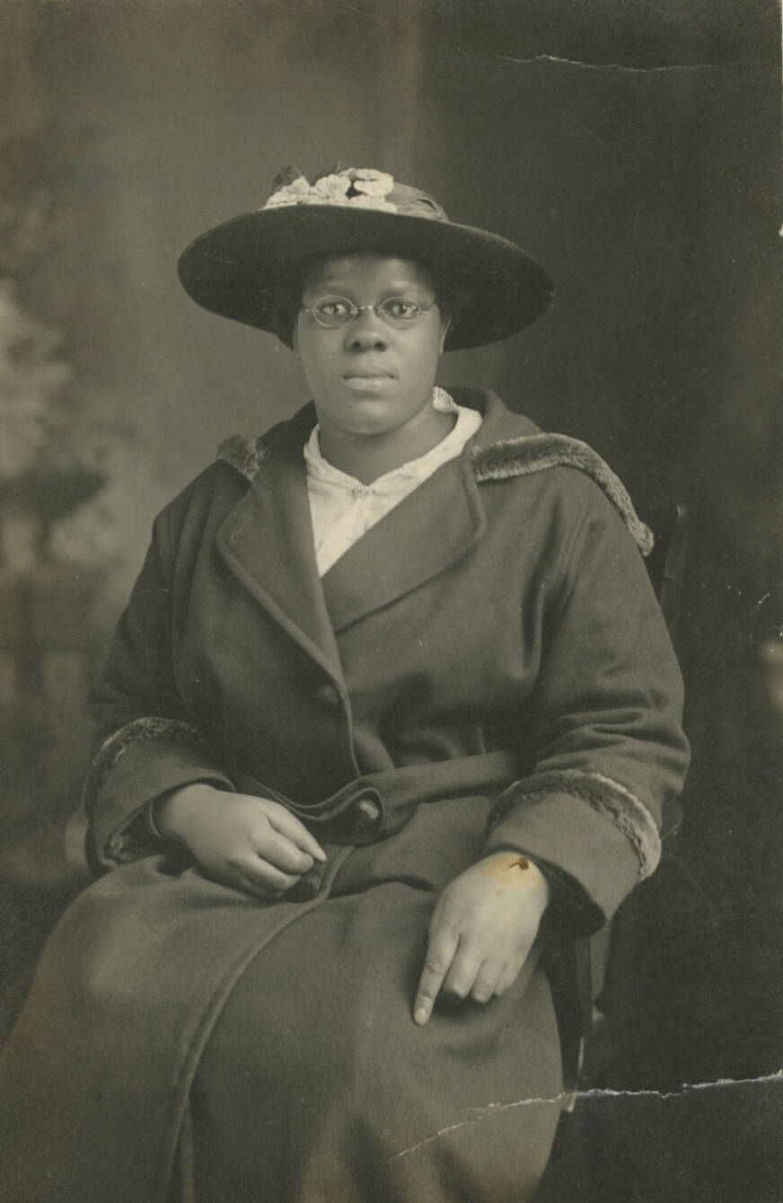 Black and white photographic portrait of a woman, seated in a chair. She wears a wide-brimmed hat with a dark jacket.